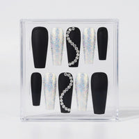 A set of coffin-shaped press-on nails displayed inside a clear acrylic case. The nails feature a black base with either solid black polish or a silver holographic glitter finish, decorated with one black nail featuring a curved line of clear rhinestones on each side.