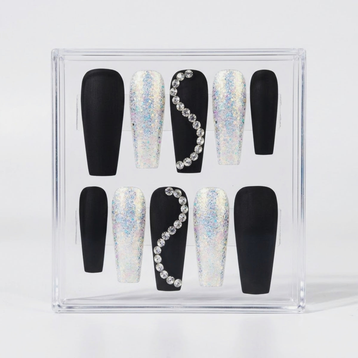 A set of coffin-shaped press-on nails displayed inside a clear acrylic case. The nails feature a black base with either solid black polish or a silver holographic glitter finish, decorated with one black nail featuring a curved line of clear rhinestones on each side.