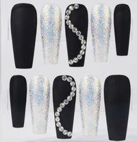 A set of coffin-shaped press-on nails displayed in a clear plastic tray, featuring matte black and white iridescent glitter designs with rhinestone accents.