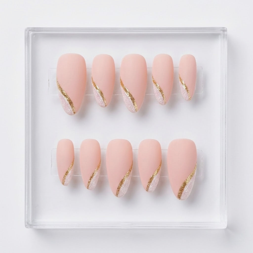 A set of ten press-on nails arranged in two rows inside a clear square tray. The nails are a soft blush pink with a diagonal metallic gold glitter accent tip.