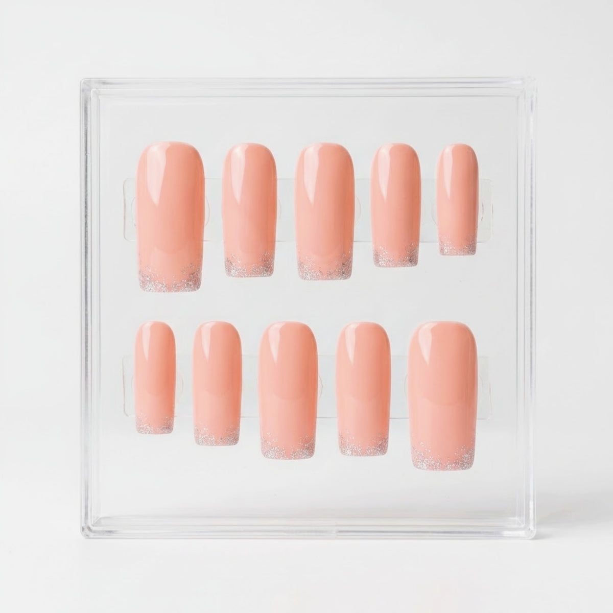 A set of ten square press-on nails in a peachy pink color with a glittery silver French tip, displayed inside a clear acrylic case on a white background.