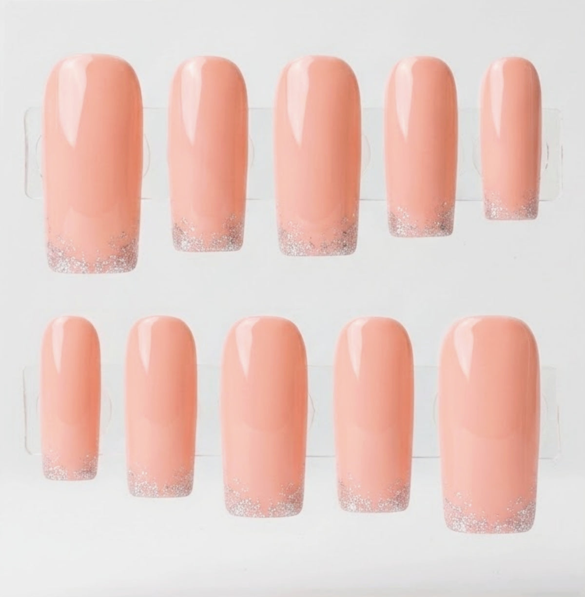 Ten square press-on nails arranged in two rows on a clear display stand against a white background. The nails have a glossy peachy pink body with a silver glitter gradient at the tips.