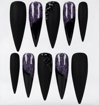 Ten stiletto-shaped press-on nails arranged in two rows on a white background. The nails are primarily black, with three featuring a glittery purple gradient design and one with black rhinestone embellishments.