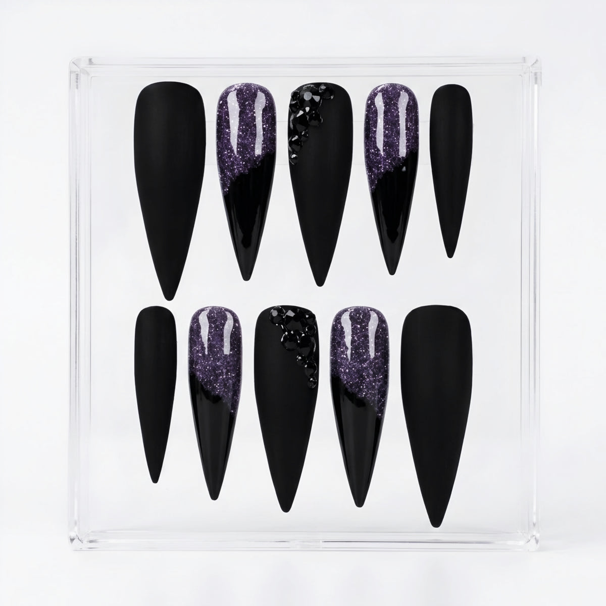 A set of ten black stiletto press-on nails in a clear plastic display box, featuring two nails with a purple glitter design and two with black glitter accents.