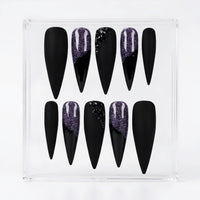 A set of ten black stiletto press-on nails in a clear plastic display box, featuring two nails with a purple glitter design and two with black glitter accents.