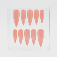 Ten stiletto-shaped press-on nails in a glossy peach-pink shade, arranged in two rows inside a clear plastic display box on a white background.