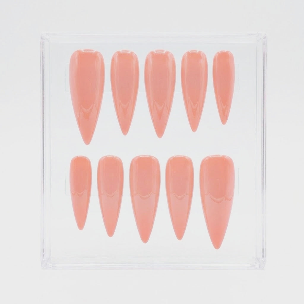 Ten stiletto-shaped press-on nails in a glossy peach-pink shade, arranged in two rows inside a clear plastic display box on a white background.