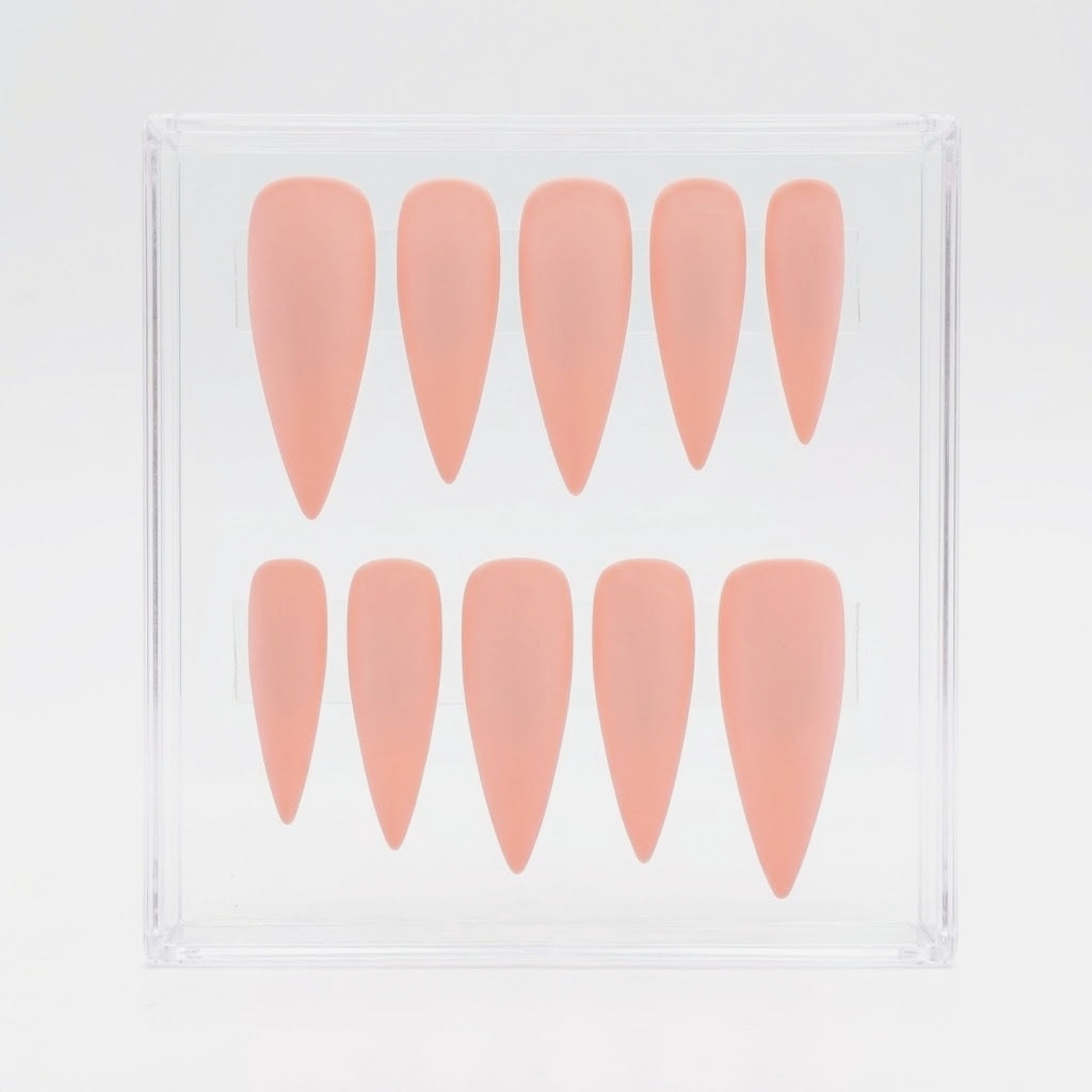 Ten stiletto-shaped press-on nails in a soft peachy pink shade, arranged in two rows inside a clear plastic display case on a white background.