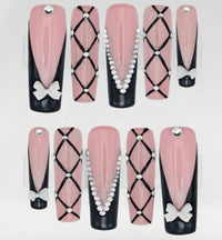 A set of press-on square nails displayed against a white background. The nails feature a glossy pink and black French manicure design with black criss-cross lattice patterns, rhinestones, and white bow accents.
