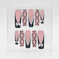 A set of 10 square press-on nails displayed in a clear acrylic case against a white background. The nails feature a pink and black color scheme with various decorative patterns including bows, rhinestones, and geometric diamond grids.
