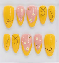 A set of almond press-on nails displayed on a white background, featuring a mix of solid bright yellow nails with black doodle line art and pastel pink nails decorated with white daisies, small yellow bumblebees, and tiny white dots.