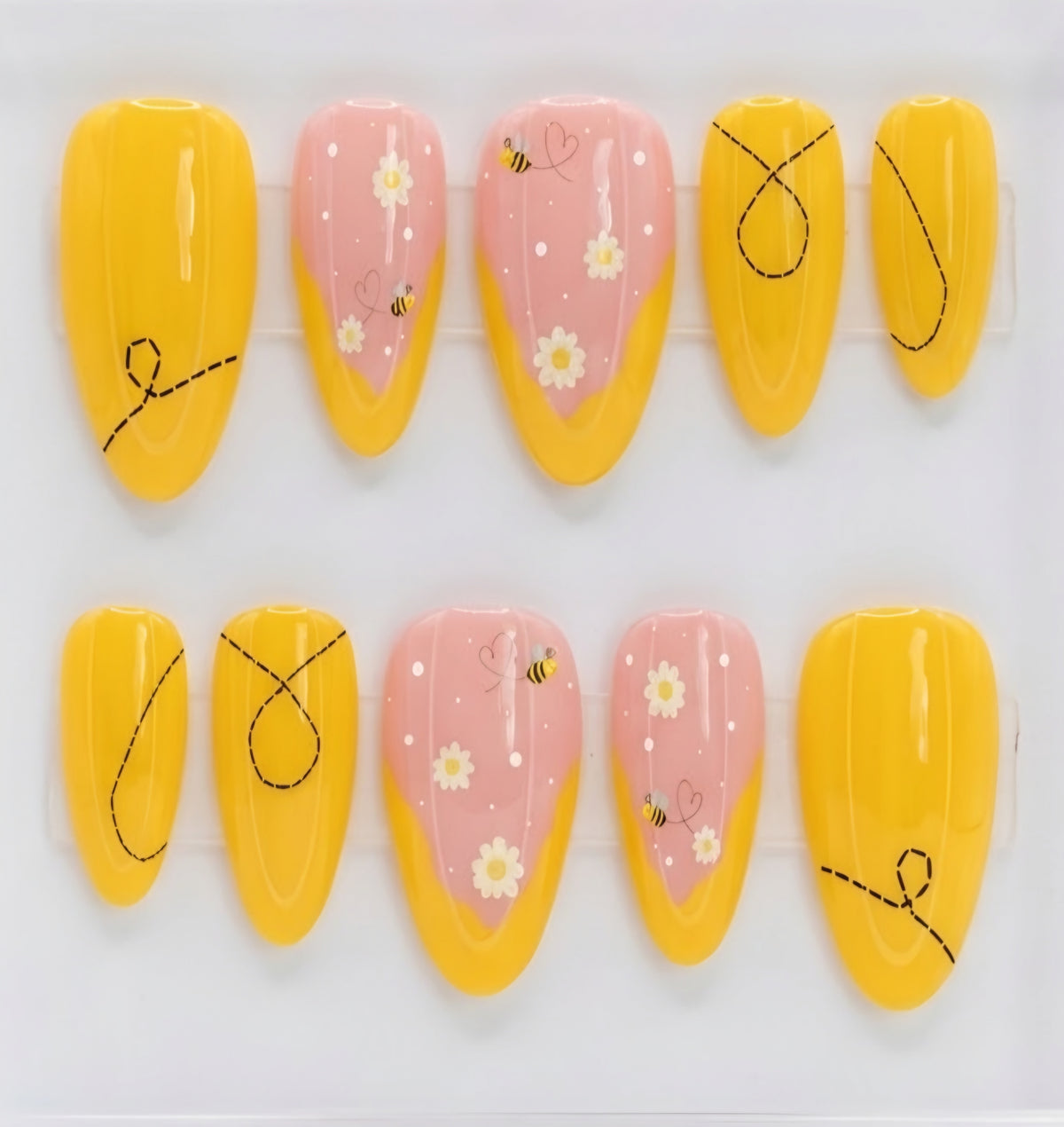 A set of almond press-on nails displayed on a white background, featuring a mix of solid bright yellow nails with black doodle line art and pastel pink nails decorated with white daisies, small yellow bumblebees, and tiny white dots.