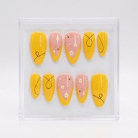 A set of almond press-on nails displayed on a white background, featuring a mix of solid bright yellow nails with black doodle line art and pastel pink nails decorated with white daisies, small yellow bumblebees, and tiny white dots.