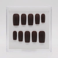 A set of ten dark brown press-on nails arranged in two rows inside a clear plastic display box on a light gray background.