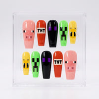 A set of 10 glossy coffin-shaped press-on nails displayed in a clear acrylic case. The nails feature pixelated Minecraft-style designs, including pink pigs, green creepers, yellow slimes, and red TNT boxes.