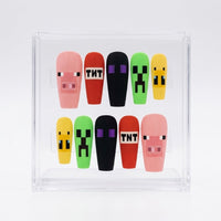 A set of mat 10 Minecraft-themed coffin shaped press on nails displayed inside a clear acrylic case on a white background. The nails feature pixelated designs including pink pigs, red TNT boxes, black ghosts, green creepers, and yellow slimes.e
