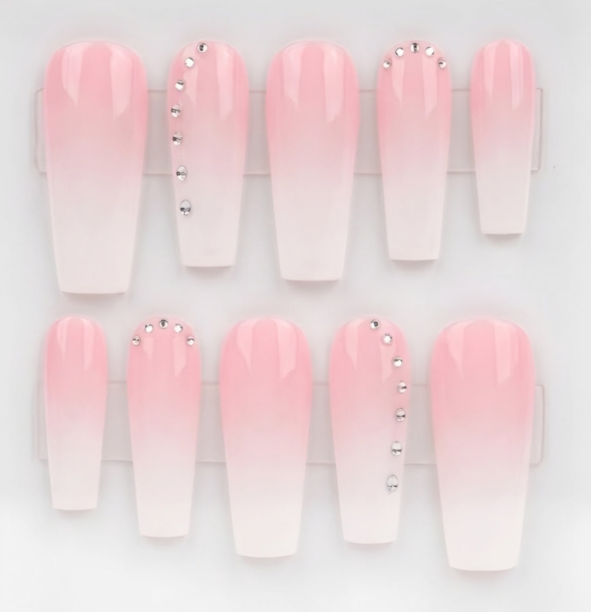 A set of coffin-shaped press-on nails with a pink-to-white ombre gradient, decorated with silver rhinestones on two of the nails, displayed in a clear plastic case against a white background.