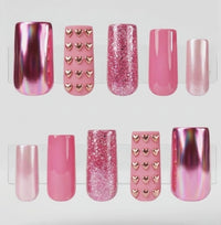 A set of 10 square press-on nails displayed on clear plastic tabs against a white background. The set includes matte pink with chrome gold hearts, glossy pink, glittery pink, chrome pink, and cat eye pink nails decorated.