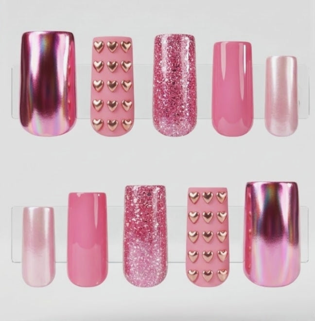A set of 10 square press-on nails displayed on clear plastic tabs against a white background. The set includes matte pink with chrome gold hearts, glossy pink, glittery pink, chrome pink, and cat eye pink nails decorated.
