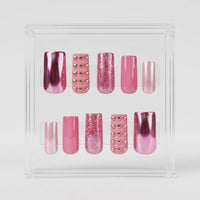 A set of 10 square press-on nails displayed on clear plastic tabs against a white background. The set includes matte pink with chrome gold hearts, glossy pink, glittery pink, chrome pink, and cat eye pink nails decorated.