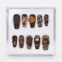 A set of 10 decorative press-on nails displayed in a clear plastic case. The nails feature various chocolate-themed designs, including a chocolate bar, ice cream cone, cookie, chocolate drizzle, and sprinkles.