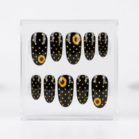 A set of ten glossy oval press-on nails displayed in a clear plastic case, featuring a black background with yellow polka dots and sunflower designs.