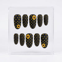 A set of ten oval matte press-on nails displayed in a clear acrylic case against a white background. The nails are black with yellow polka dots and two yellow sunflower designs.