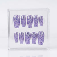 A set of ten glittery lavender-purple press-on nails displayed inside a clear acrylic box against a white background.
