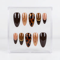 A set of ten press-on nails displayed in a clear acrylic case against a white background. The nails feature various designs in shades of brown, beige, and black, including solid dark brown, nude beige, leopard print, and marbled patterns with thin gold accent lines.