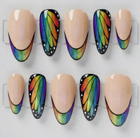 Ten almond press-on nails displayed on clear plastic hangers, featuring a mix of solid nude beige nails and butterfly-wing style nails with a black base, white dots, and a vibrant rainbow gradient.