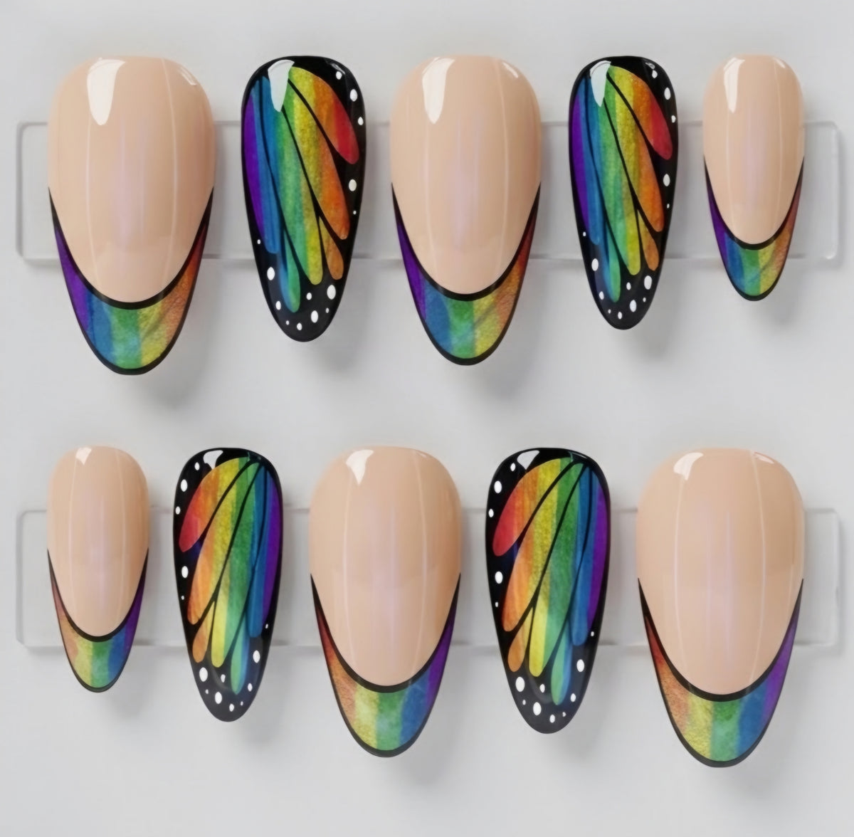 Ten almond press-on nails displayed on clear plastic hangers, featuring a mix of solid nude beige nails and butterfly-wing style nails with a black base, white dots, and a vibrant rainbow gradient.