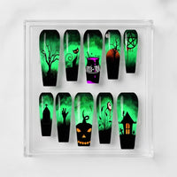 A set of coffin-shaped press on nails displayed in a clear acrylic case, featuring spooky Halloween designs like a tree, a cat, a potion bottle, a tombstone, a jack-o'-lantern, and a haunted house over a glowing green misty background. 