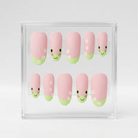 A set of 10 oval matte pink press-on nails with a cute green frog design, small white flowers, displayed inside a clear acrylic case on a light gray background.