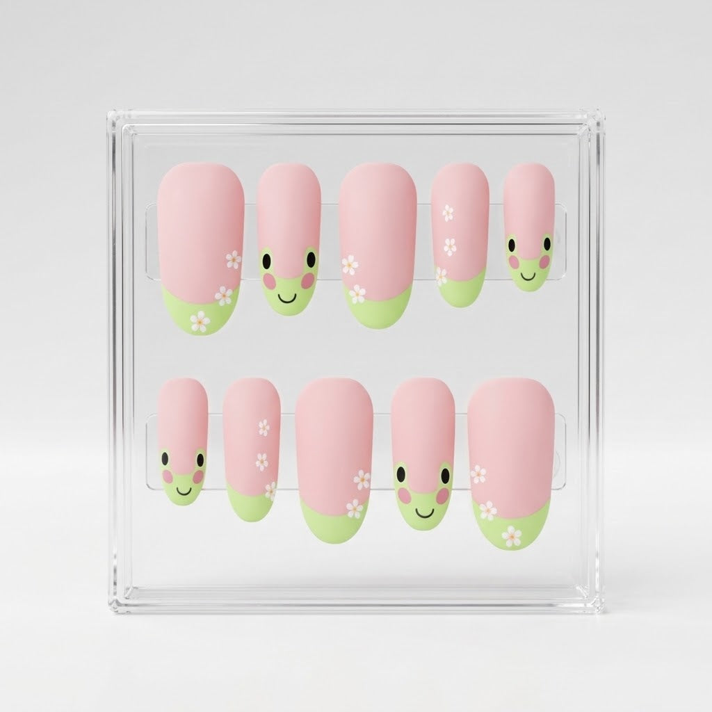 A set of 10 oval matte pink press-on nails with a cute green frog design, small white flowers, displayed inside a clear acrylic case on a light gray background.