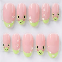 A set of 10 oval glossy pink press-on nails with a cute green frog design, small white flowers, displayed inside a clear acrylic case on a light gray background.