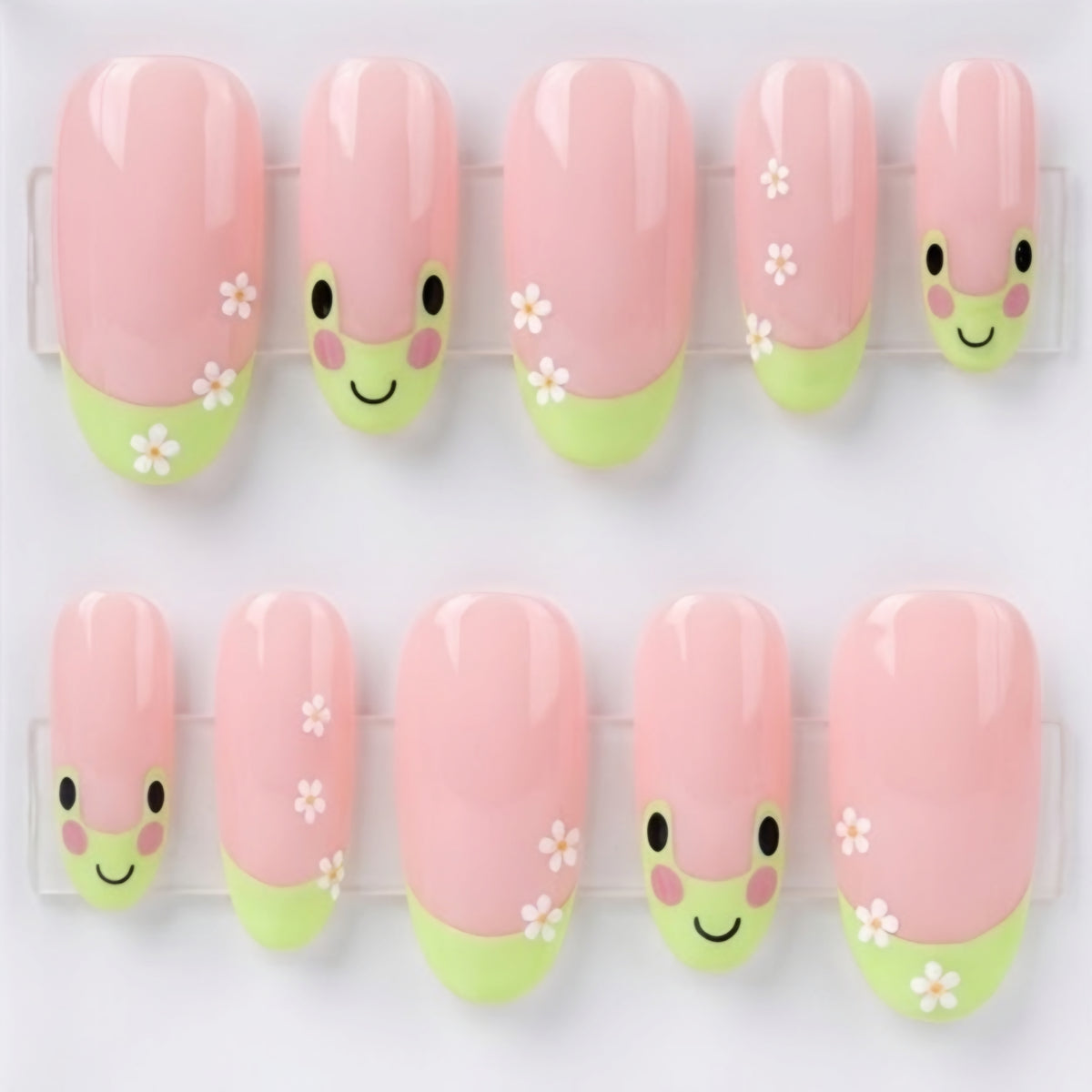 A set of 10 oval glossy pink press-on nails with a cute green frog design, small white flowers, displayed inside a clear acrylic case on a light gray background.