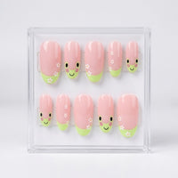 A set of 10 oval glossy pink press-on nails with a cute green frog design, small white flowers, displayed inside a clear acrylic case on a light gray background.