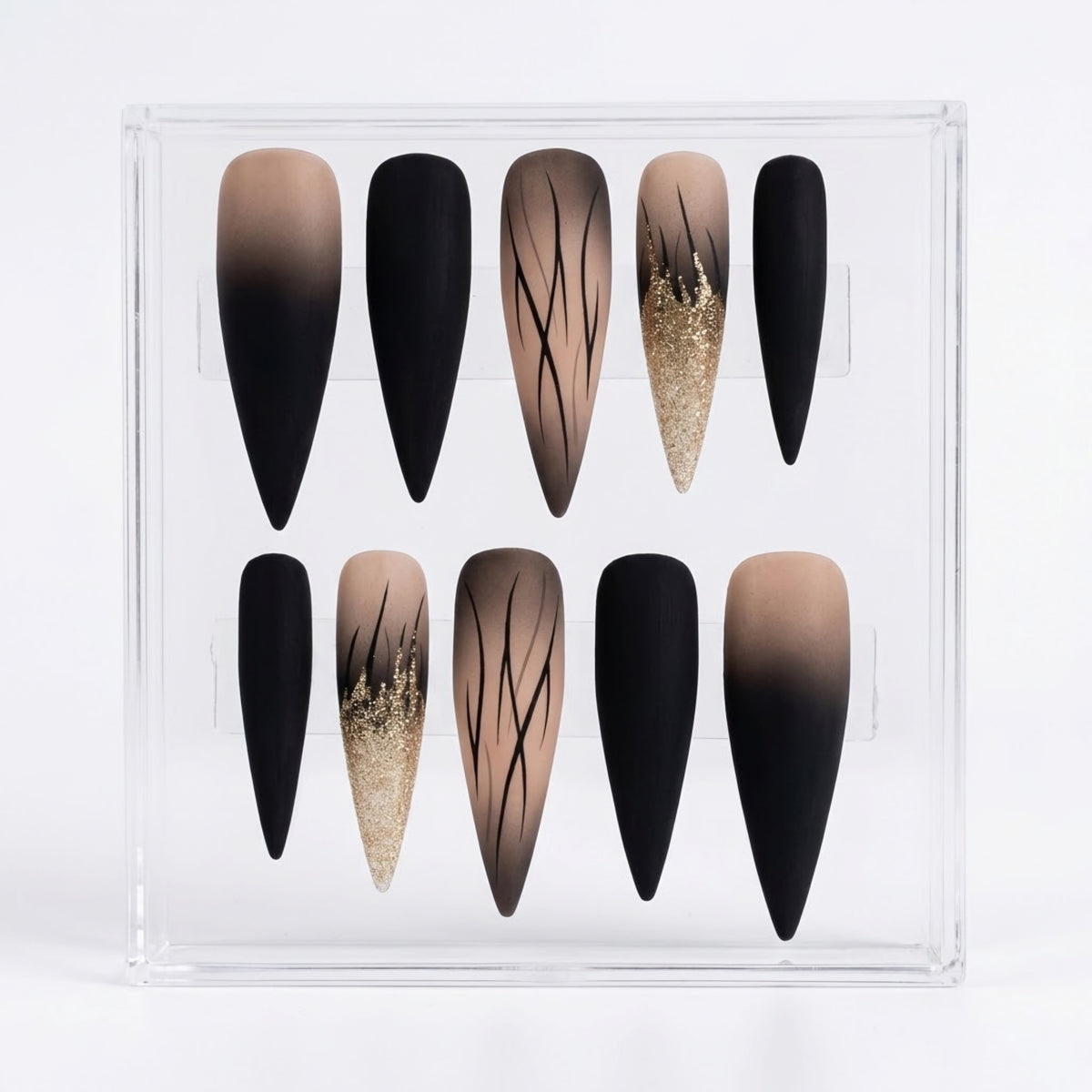 A set of 10 long stiletto-shaped press-on nails displayed in a clear acrylic case against a white background. The nails feature a mix of solid black, beige-to-black ombre gradients, and a textured gold glitter design with thin black branch-like lines.