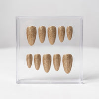 Ten almond-shaped press-on nails with a textured gold glitter finish, displayed inside a clear plastic storage case on a light gray background.