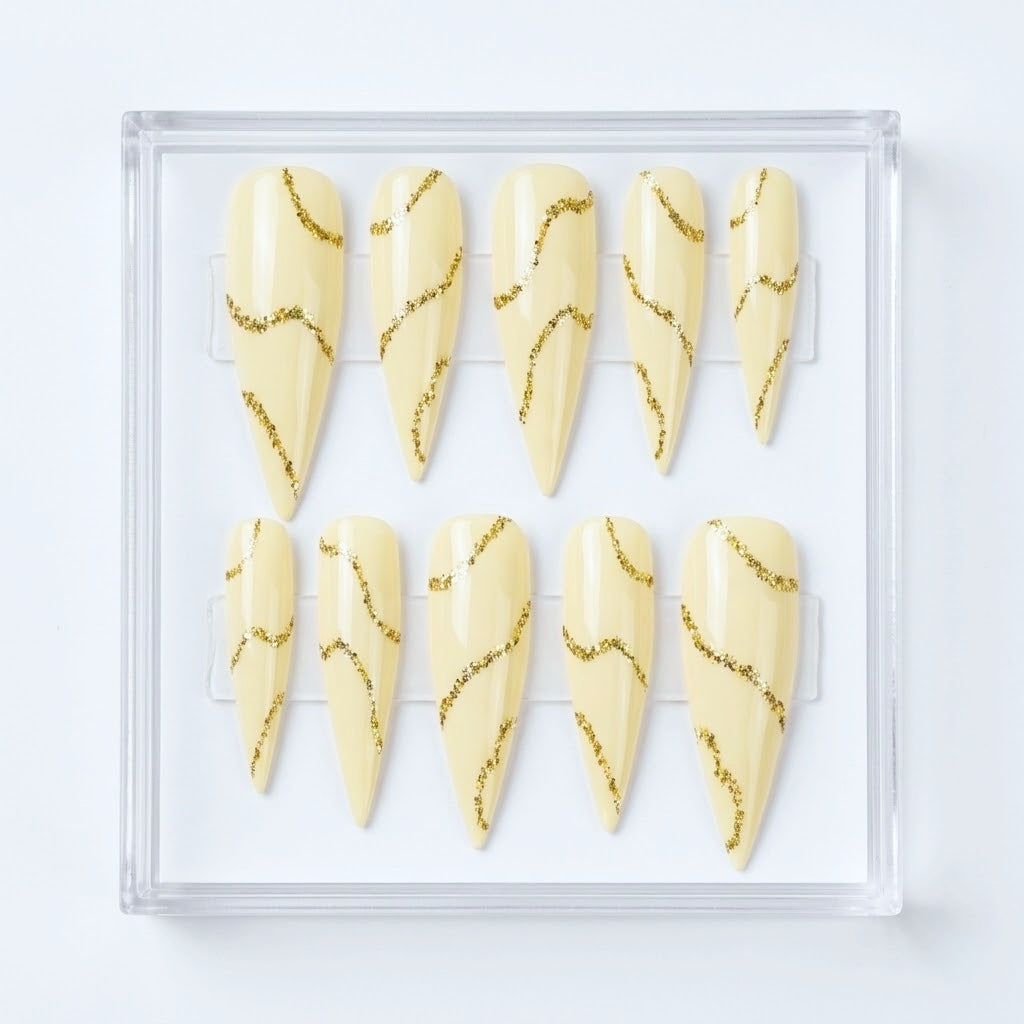 Ten stiletto-shaped press-on nails in a clear plastic case, featuring a glossy  cream base with wavy metallic gold glitter lines.