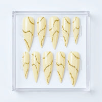 Ten stiletto-shaped press-on nails in a clear plastic case, featuring a glossy  cream base with wavy metallic gold glitter lines.