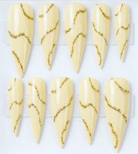 Ten stiletto-shaped press-on nails with a glossy pale cream base and wavy metallic gold glitter lines, arranged on a clear display card against a white background.