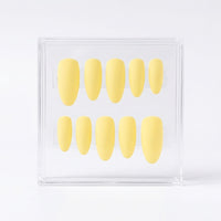 A set of ten pale yellow press-on nails arranged in two rows inside a clear acrylic display box on a white background.