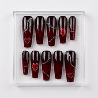A set of coffin-shaped glossy press-on nails in a clear plastic display box. The nails have a glossy deep red-to-black ombre gradient with silver barbed wire details and red heart designs.