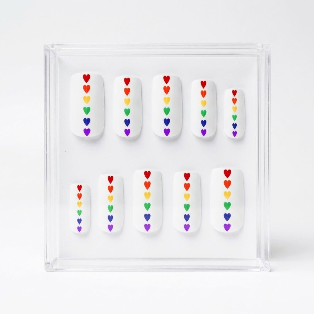 Set of 10 square matte white press on nails with small rainbow hearts running down the centre. Displayed in a  clear display case on a white background.
