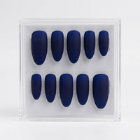 A set of ten matte dark blue press-on nails arranged in two rows inside a clear plastic display box on a light gray background.