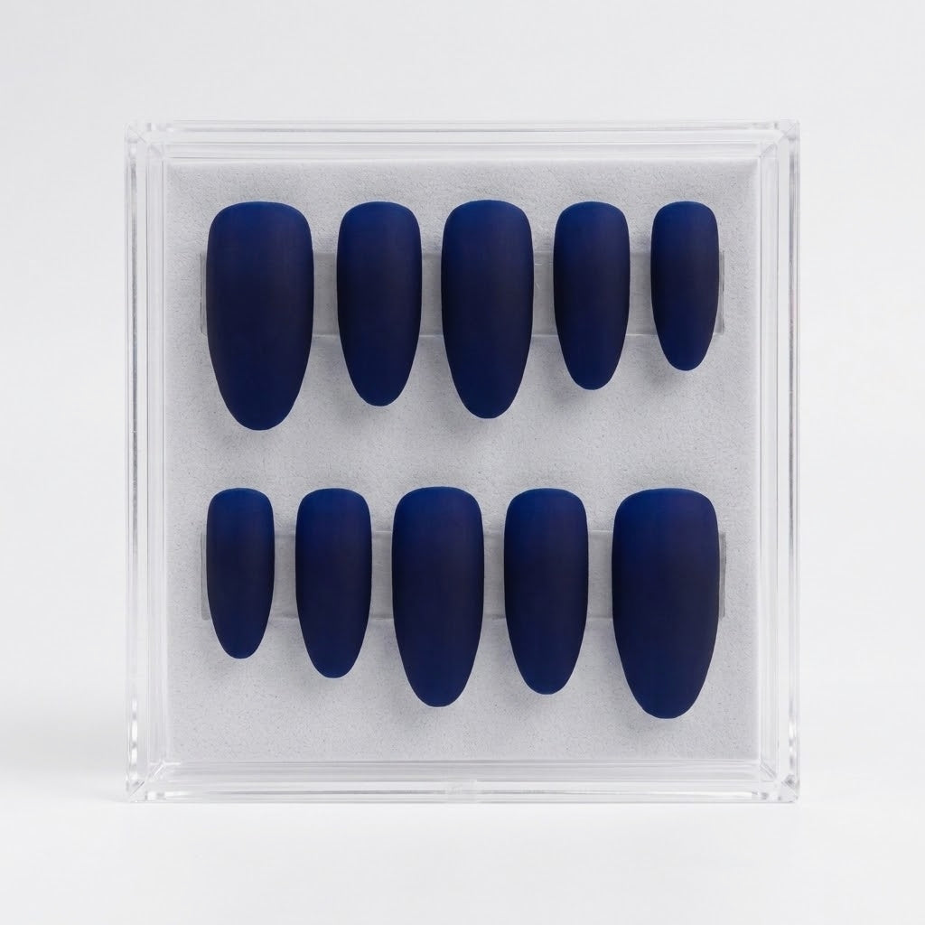 A set of ten matte dark blue press-on nails arranged in two rows inside a clear plastic display box on a light gray background.