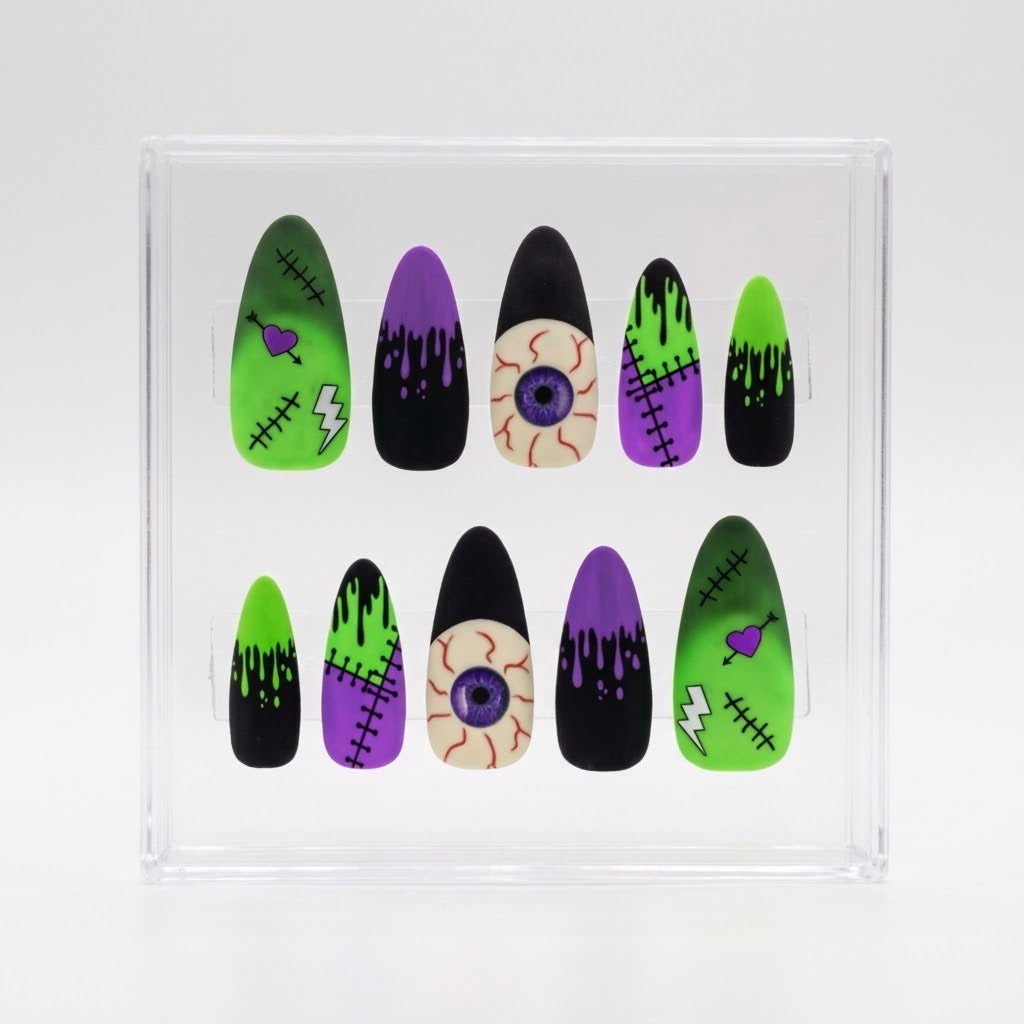 A set of Halloween-themed press-on nails displayed inside a clear plastic case on a white background. The nails feature designs inspired by Frankenstein, including green and purple stitched heads, black and purple dripping monsters, a stitched lightning bolt nail, and a realistic eyeball nail.