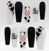 A set of coffin-shaped press-on nails displayed on clear stands, featuring a mix of solid black, white, and cow-print designs with one nail showing the word 'Moo' in script.