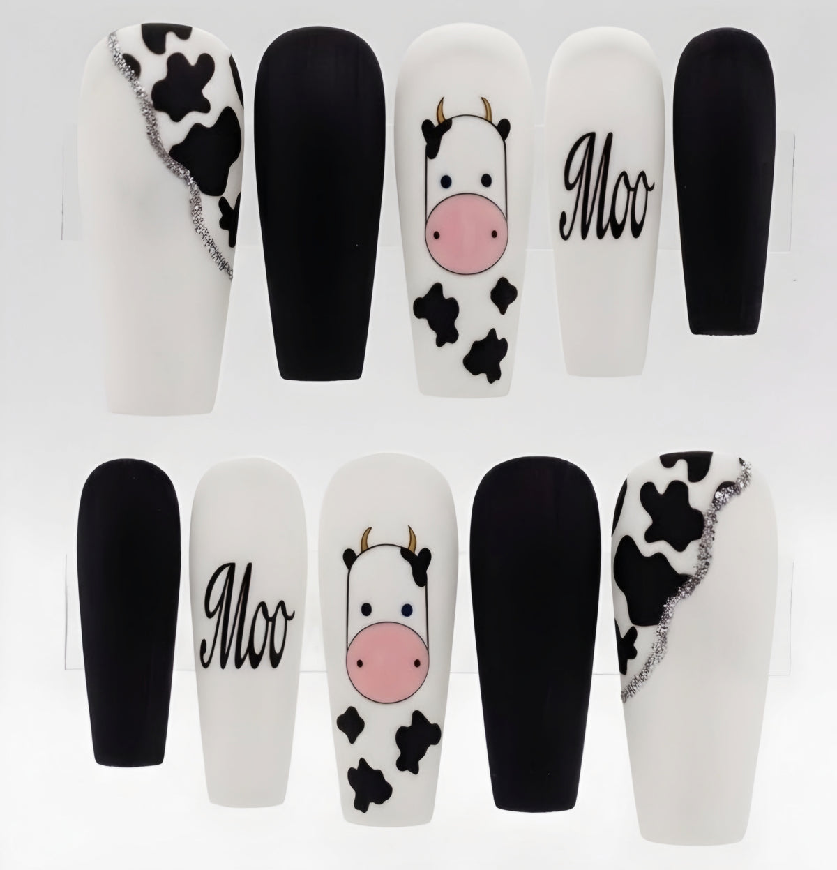 A set of coffin-shaped press-on nails displayed on clear stands, featuring a mix of solid black, white, and cow-print designs with one nail showing the word 'Moo' in script.